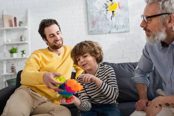 Junge zeigt auf Bauklötze neben lächelndem Vater und Großvater auf Couch — Stockfoto