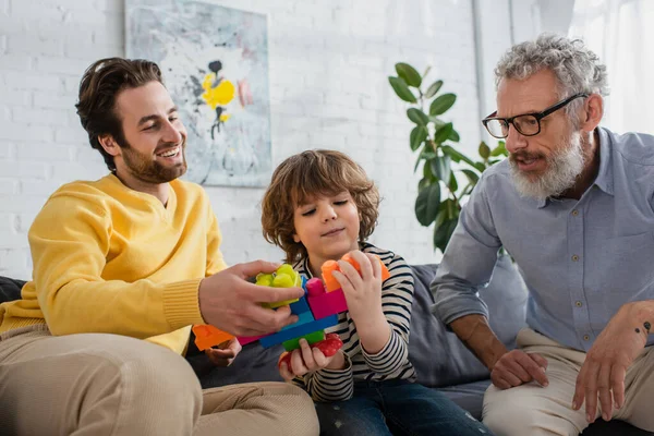 Lächelnder Vater spielt mit Sohn neben Großvater auf Couch Bauklötze — Stockfoto