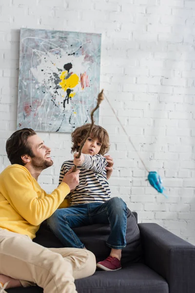 Pai abraçando criança com vara de pesca de brinquedo na sala de estar — Fotografia de Stock