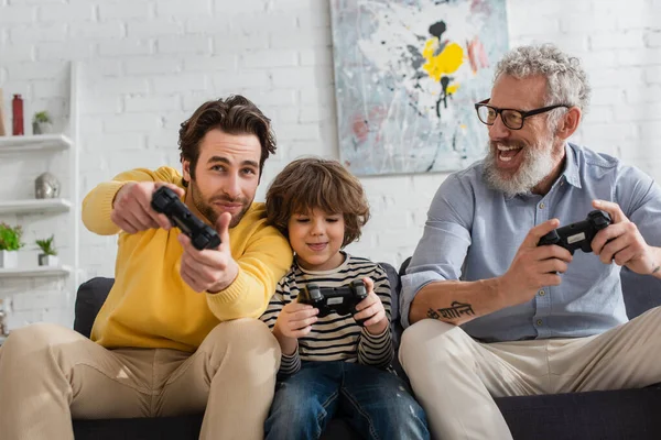 KYIV, UCRANIA - 12 de abril de 2021: Joven jugando videojuegos con su hijo y su padre en el sofá - foto de stock