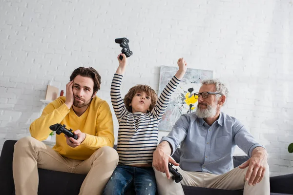 KYIV, UCRANIA - 12 DE ABRIL DE 2021: Vista de ángulo bajo del niño emocionado con joystick cerca de padres tristes en el sofá - foto de stock