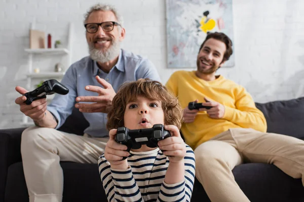 KYIV, UCRANIA - 12 DE ABRIL DE 2021: Un niño sorprendido jugando un videojuego con un padre y un abuelo borrosos - foto de stock