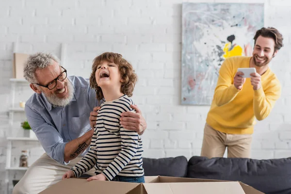 Großvater umarmt Kind in Schachtel neben verschwommenem Vater mit Smartphone — Stockfoto