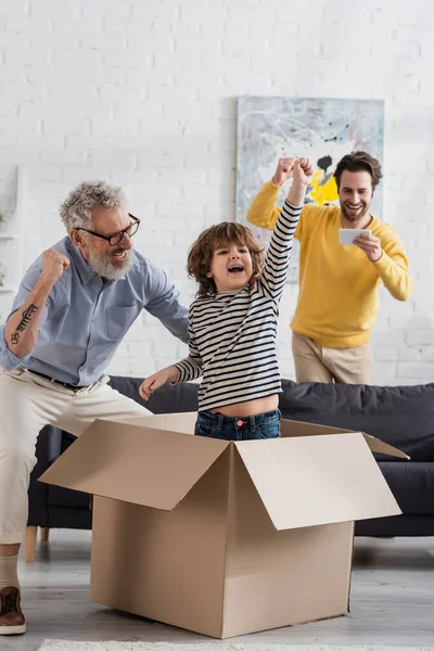 Excited kid showing yes gesture in carton box near parent with smartphone and grandfather — Stock Photo