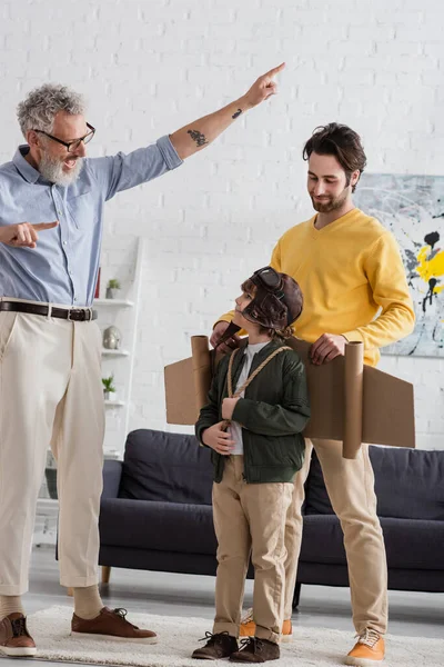 Alegre abuelo señalando con los dedos cerca de niño en traje de aviador e hijo - foto de stock