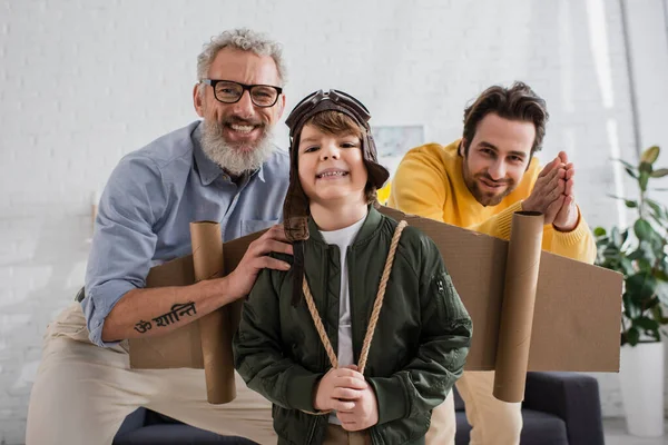 Sonriente niño en traje de aviador mirando a la cámara cerca del abuelo y el padre - foto de stock