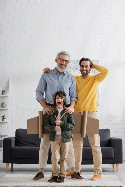 Lächelnde Männer stehen neben Kind im Fliegerkostüm zu Hause — Stockfoto