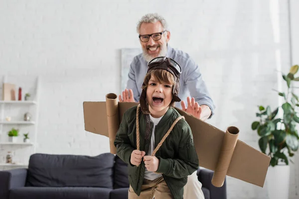 Excited boy in aviator costume playing near blurred granddad — Stock Photo