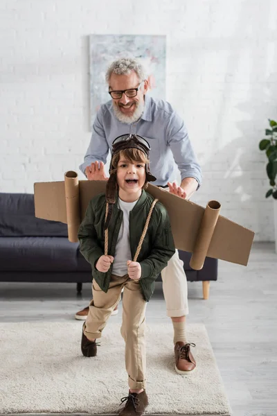 Niño feliz en casco de aviador y alas jugando con el abuelo - foto de stock