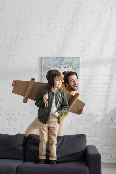 Smiling father standing near son in aviator costume on couch — Stock Photo