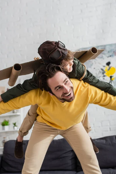 Smiling father playing with son in aviator goggles and carton wings — Stock Photo