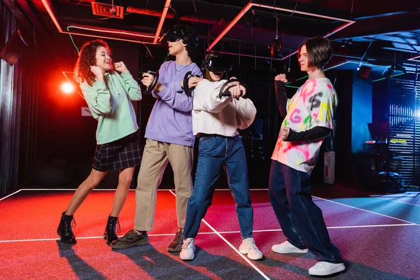 Adolescentes interracial emocionados mostrando el gesto de ganar cerca de los amigos jugando en auriculares vr - foto de stock