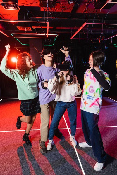 Excitada chica afroamericana mostrando gesto de victoria cerca de amigos jugando en auriculares vr - foto de stock