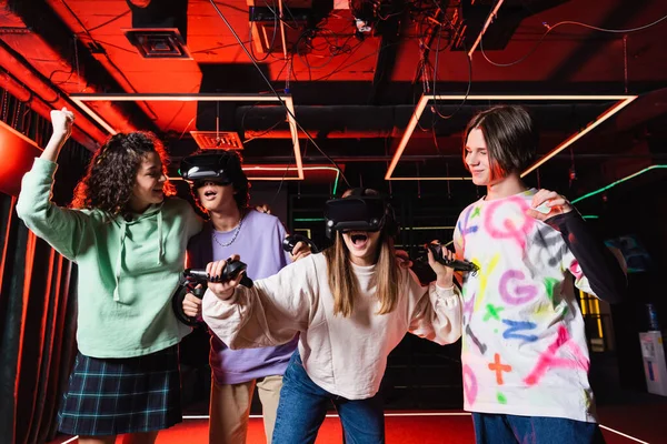 Afroamericana adolescente mostrando el gesto de triunfo cerca de amigos sorprendidos en auriculares vr - foto de stock