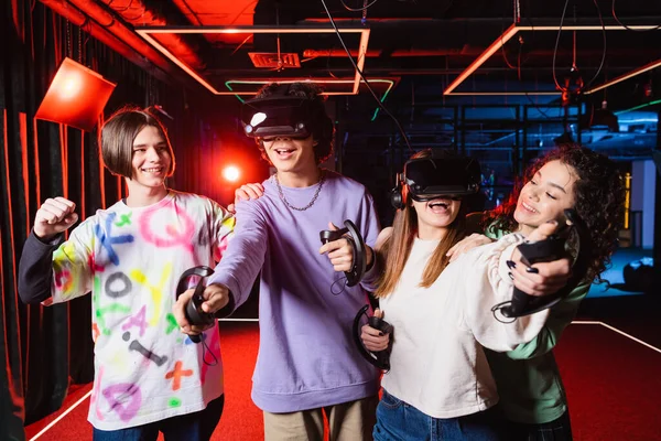Smiling teenager showing win gesture near multiethnic friends in vr headsets — Stock Photo