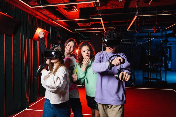 Asombrado afroamericano chica cubriendo la boca con la mano cerca de amigos en vr sala de juegos - foto de stock