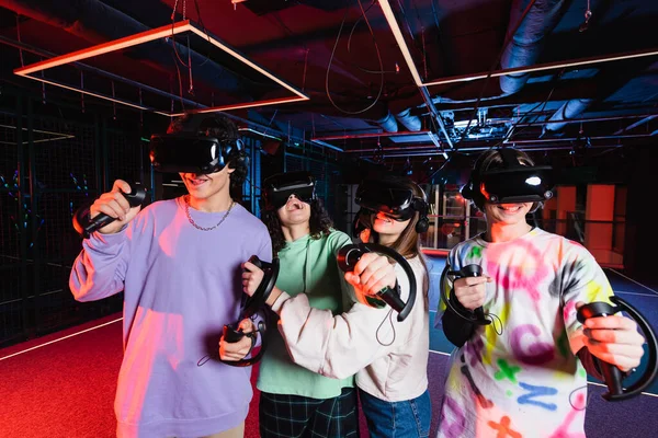 Возбужденные многонациональные друзья-подростки в виртуальных наушниках игры в игровой комнате — стоковое фото