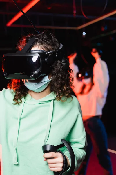 Filles interracial dans des masques médicaux et des casques vr jeu dans la zone de jeu sur fond flou — Photo de stock