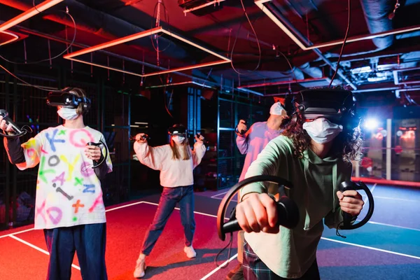 Мультиэтнические друзья в медицинских масках и VR-наушниках развлекаются в игровой комнате — стоковое фото