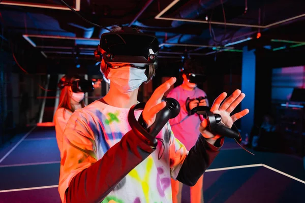 Adolescent garçon dans masque médical et vr casque de jeu près des amis flous — Photo de stock