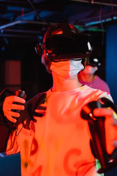 Teenager-Gamer mit medizinischer Maske und vr-Headset in der Nähe von Freund auf verschwommenem Hintergrund — Stockfoto