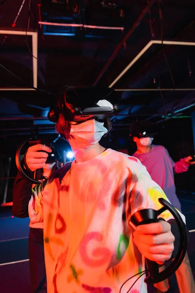 Мальчик-подросток в медицинской маске игры вблизи размытых друзей в виртуальной игровой зоне — стоковое фото
