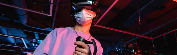 Парень-подросток в медицинской маске и видеогарнитуре игры в игровой зоне, баннер — стоковое фото