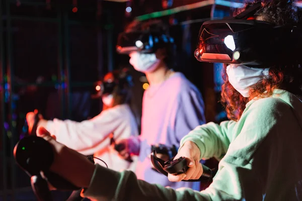 African american girl in medical mask and vr headset gaming near blurred friends — Stock Photo