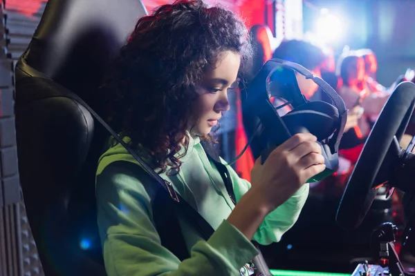 Africano americano ragazza holding vr auricolare su auto simulatore vicino offuscata amici — Foto stock