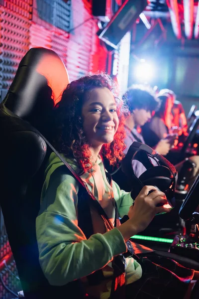 Cheerful africam american girl in car simulator smiling at camera near blurred friends — Stock Photo