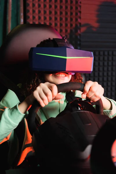 Excited african american girl in vr headset holding steering wheel of car simulator — Stock Photo