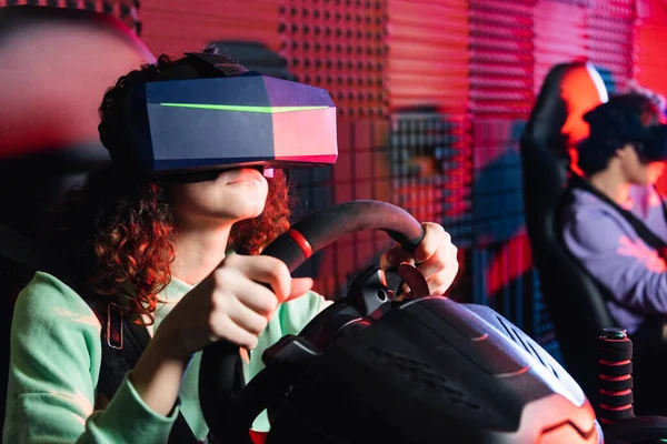 African american girl in vr headset gaming on car simulator near blurred friend — Stock Photo
