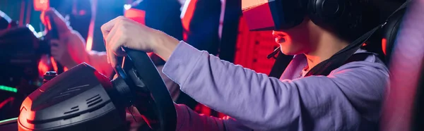 Vista parcial de adolescente en el juego de auriculares vr en simulador de coche cerca de amigo borroso, bandera - foto de stock