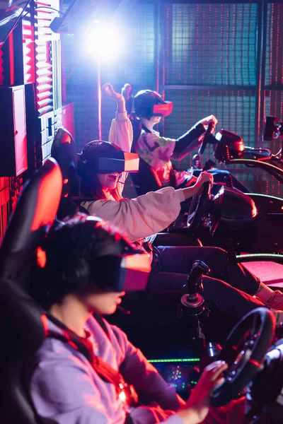 Fille excitée montrant geste de victoire près des amis flous sur les simulateurs de course automobile — Photo de stock