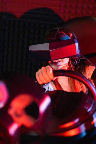 Fille souriante dans vr casque de course dans le simulateur de voiture sur le premier plan flou — Photo de stock