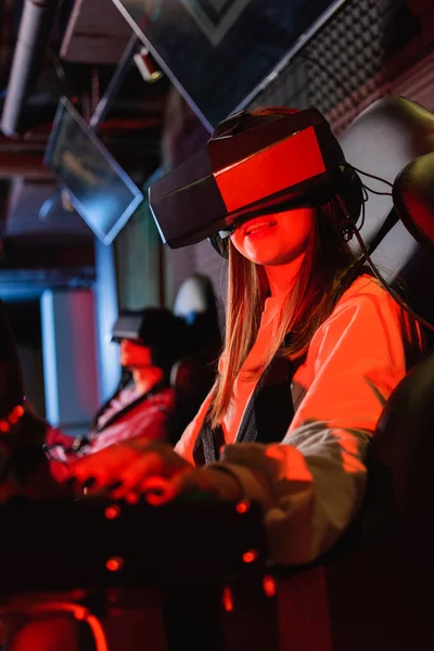 Smiling girl in vr headset gaming on car racing simulator, blurred foreground — Stock Photo