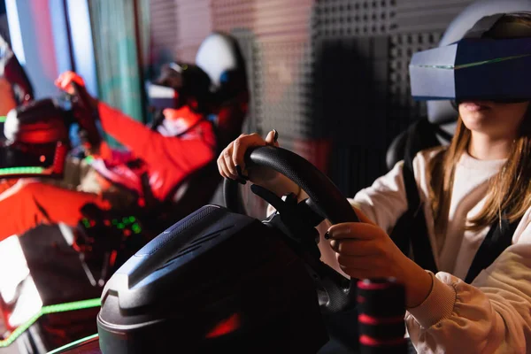 Friends in vr headsets playing racing game on car simulators, blurred foreground — Stock Photo
