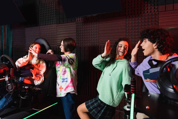 Cheerful multicultural friends giving high five near car racing simulators — Stock Photo