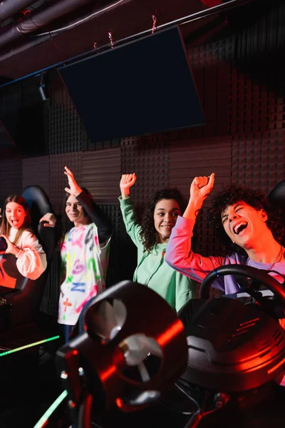 Excited multiethnic friends showing rejoice gesture near racing simulators — Stock Photo