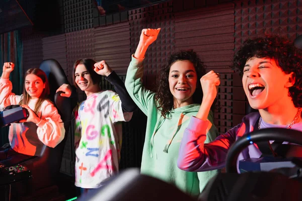 Heureux adolescents multiethniques montrant geste de victoire près de simulateurs de voiture — Photo de stock