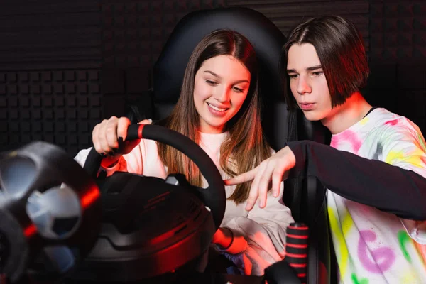 Adolescente apuntando al volante del simulador de coche cerca de un amigo sonriente - foto de stock