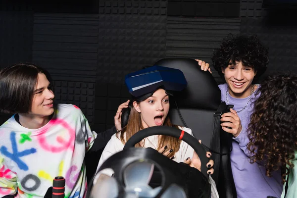 Amigos multiétnicos riendo cerca de chica asombrada en simulador de carreras de coches - foto de stock