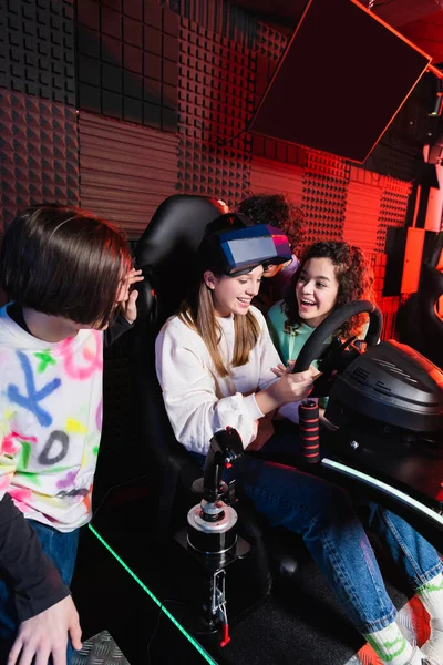 Cheerful interracial teenagers laughing near amazed girl on car simulator — Stock Photo