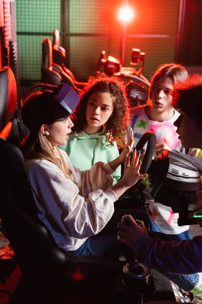 Emocionante menina gestos no carro corrida simulador perto interracial amigos — Fotografia de Stock