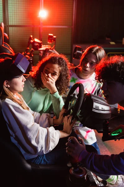 Shocked african american girl covering mouth with hand near friend on car simulator — Stock Photo