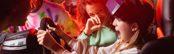 Multiethnic teens laughing near scared girl in car simulator, banner — Stock Photo