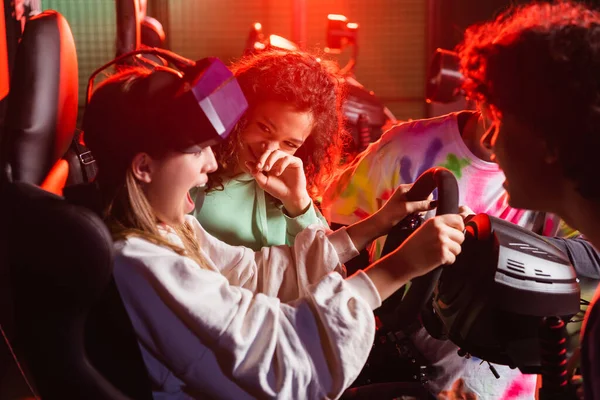 Scared teenage girl screaming on car simulator near laughing interracial friends — Stock Photo