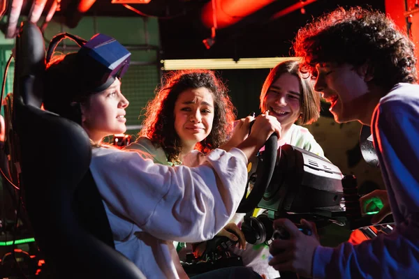 Adolescentes multiculturales riendo cerca de tensa chica de juego en simulador de coche - foto de stock