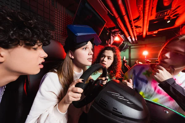 Adolescente menina jogos no carro corrida simulador perto preocupado multiétnico amigos — Fotografia de Stock