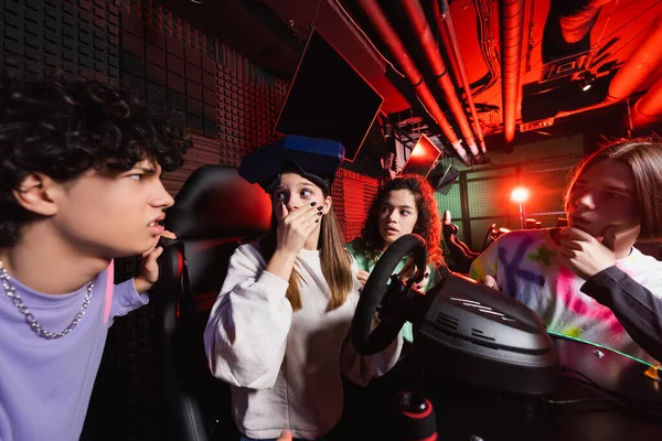 Shocked girl on racing simulator covering mouth with hand near interracial friends — Stock Photo